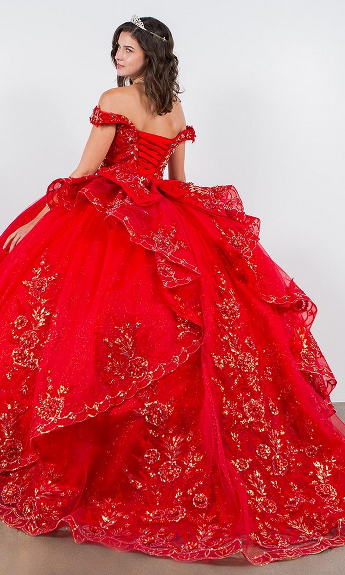 A woman wearing the Quinceanera Ball Gown 8100J, featuring gold floral embroidery, iridescent beading, and an off-the-shoulder design with a tiara, stands facing away from the camera.
